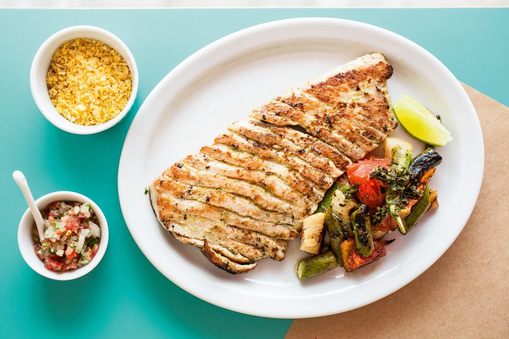 Na imagem com fundo azul, aparecem o prato barriga de atum servida com vegetais tostados. Ao lado aparecem dois potinhos com farofa e vinagrete