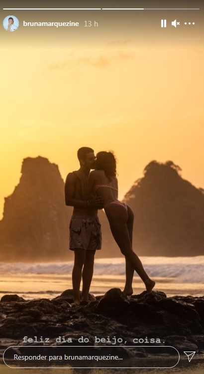 Com uma luz de entardecer, a foto exibe a silhueta de Bruna Marquezine se inclinando para beijar Enzo Celulari, ambos em pé e com uma paisagem de praia ao fundo