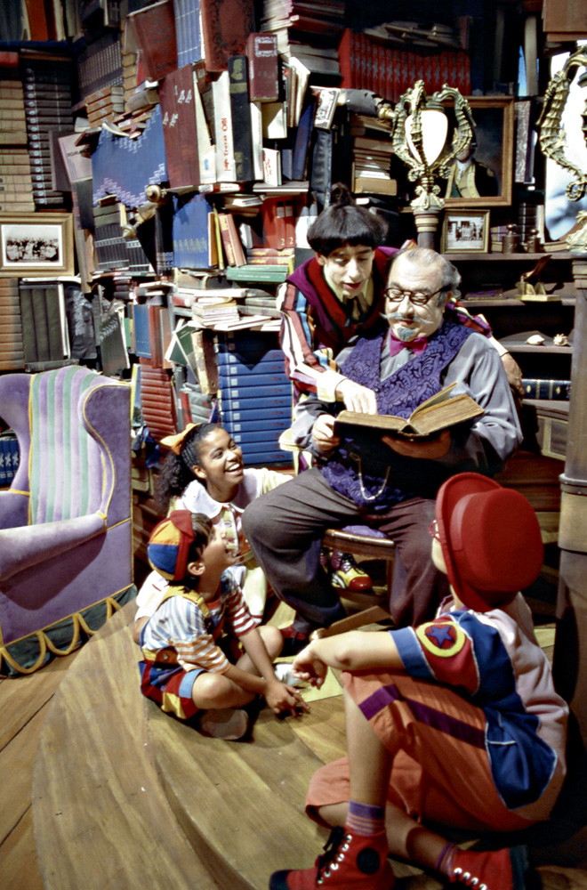 Sergio está sentando em um poltrona, com três crianças sentadas em sua frente. Atrás dele, de pé, o personagem Nino aponta para o livro que Sergio segura.