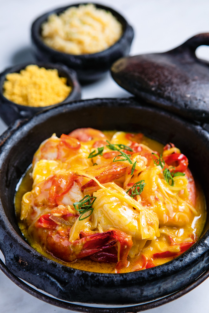 Moqueca de camarão na panela de barro e ao fundo porções de farofa e arroz