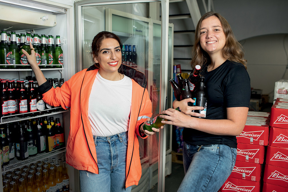 Mariam e Muriel, criadoras da startup Avocado, vendida para o Rappi, posam ao lado de geladeira lotada de cervejas. Mariam veste jaqueta laranja e Muriel, que segura várias cervejas no colo, veste camiseta preta.