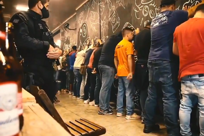 A imagem mostra pessoas com as mãos na parede em um bar durante operação policial