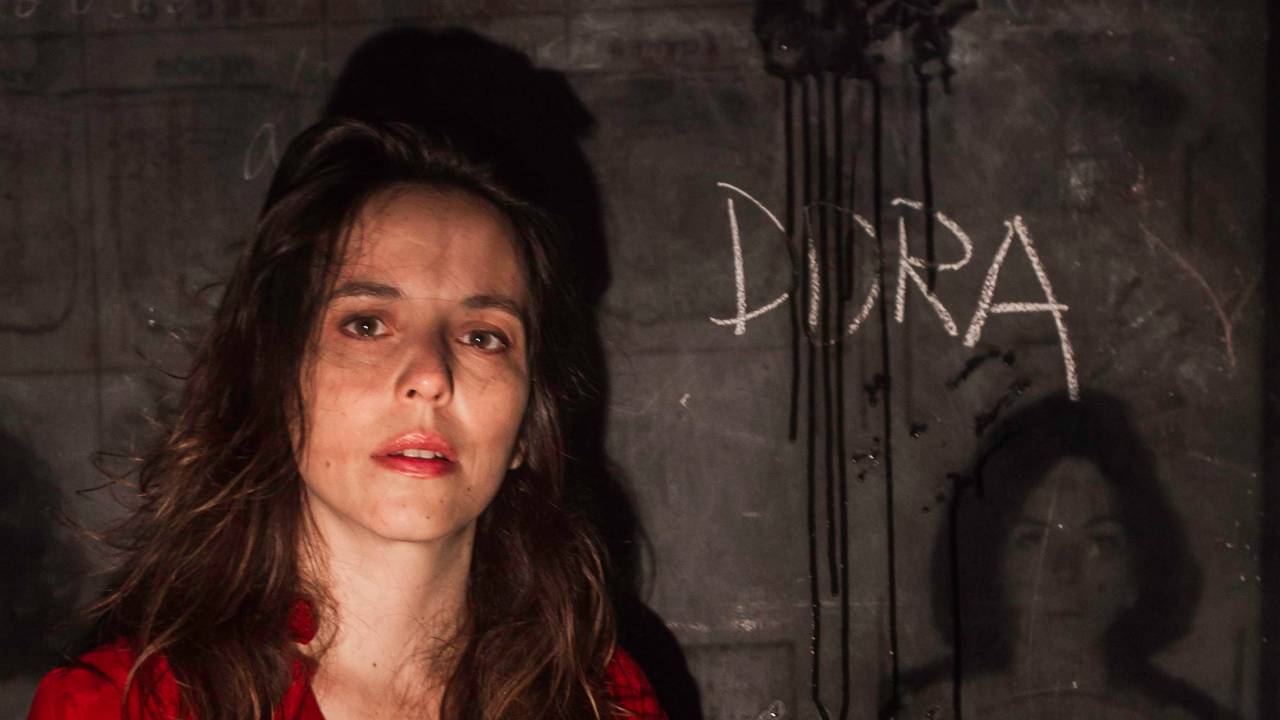 Mulher olha para a câmera com expressão de preocupação em cenário escuro e cinza. Na parede, escritos como "mamãe," e "Dora"