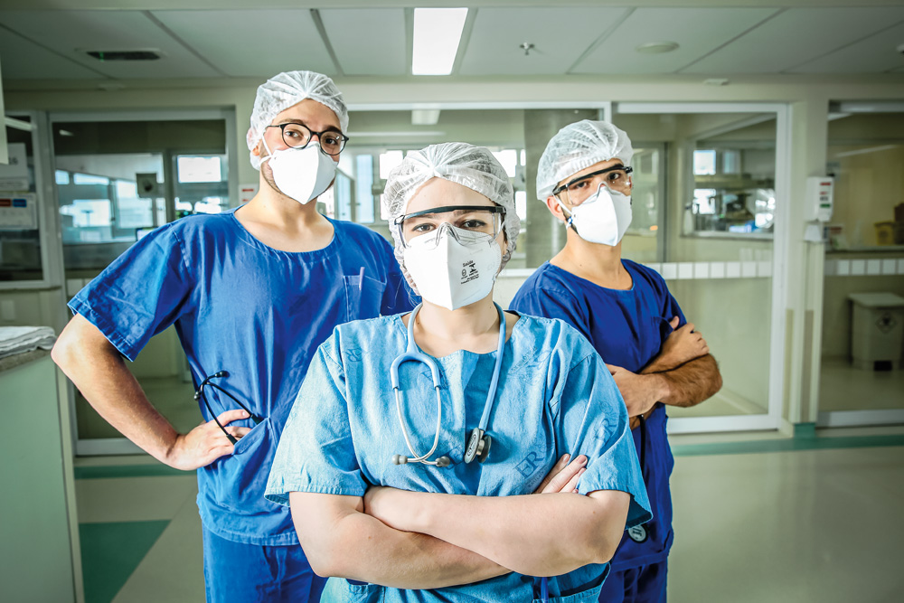 Três médicos, com roupa azul, toca, óculos de proteção e máscara, encarando a câmera