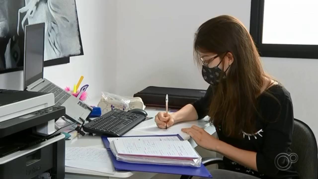 Elisa sentada em uma cadeira e escrevendo em um caderno em uma mesa em sua frente. Ela está de máscara e óculos, estudante, e ainda há mais um notebook em sua frente