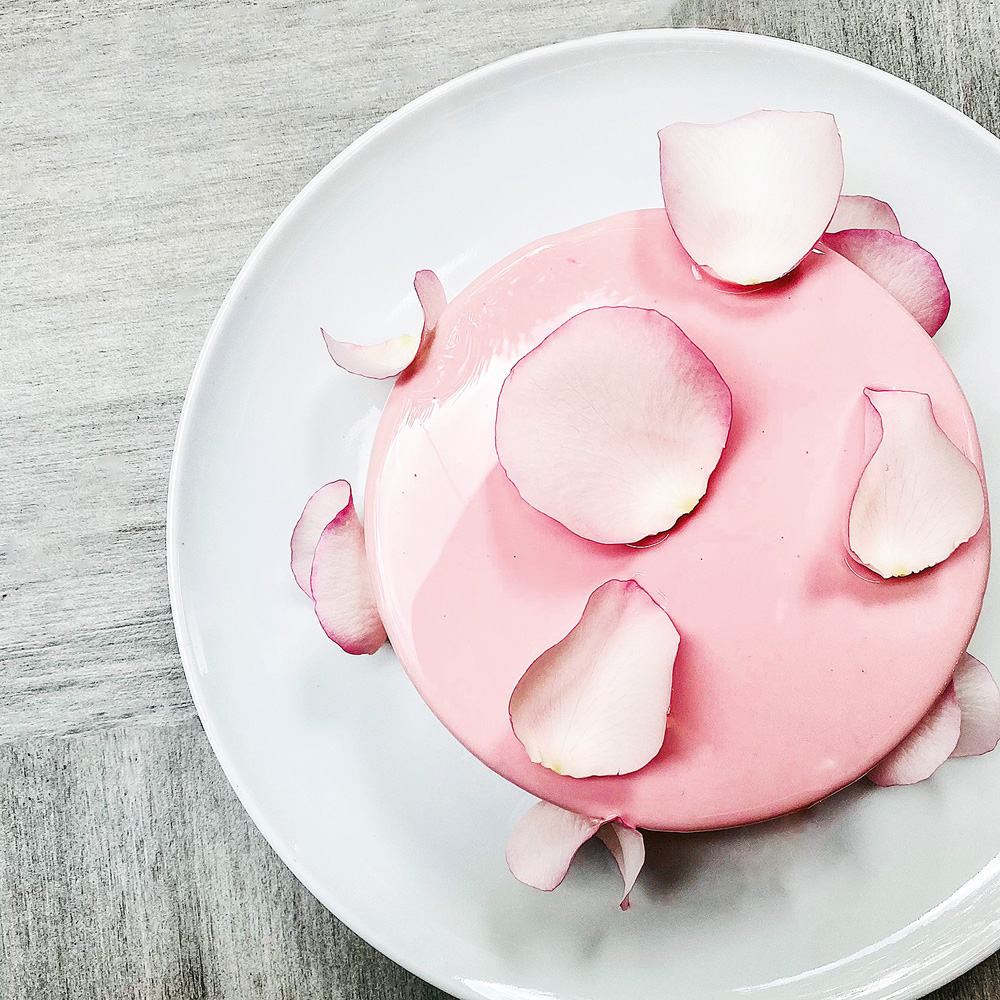 Foto tirada de cima de um bolo redondo rosa bebê brilhante com "pétalas" de rosa branca e rosa feitas de açúcar espalhadas pelo doce, que está sobre prato branco circular.