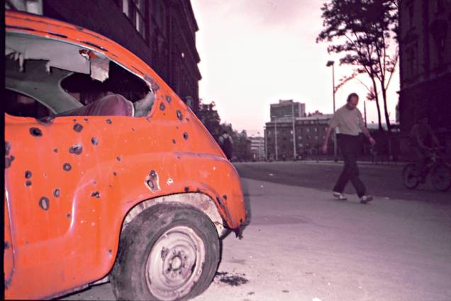 Marcas de bombardeios nas paredes e carro destruído abandonado na rua: cidade-ruína e ecos da violência