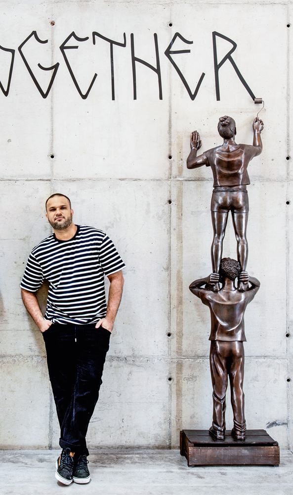 O artista Flávio Cerqueira posa ao lado de sua escultura, que mostra duas crianças em bronze, um no ombro do outro, pintando uma parede