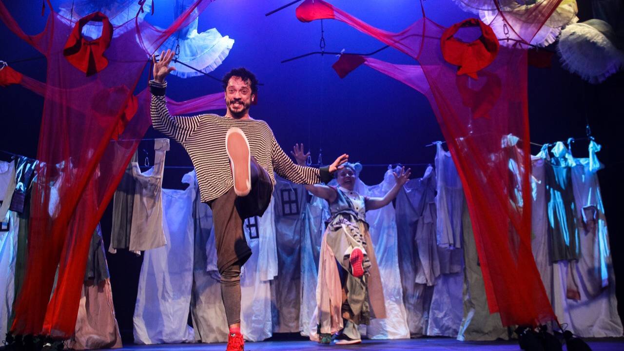 Peça de teatro mostra homem com um pé levantado e sorrindo, em um cenário lúdico com panos pendurados. Atrás dele uma moça com braços levantados