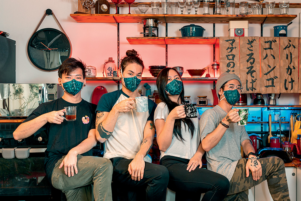 quatro pessoas agachadas com o rosto coberto por máscaras protetoras segurando canecas dentro de uma cozinha