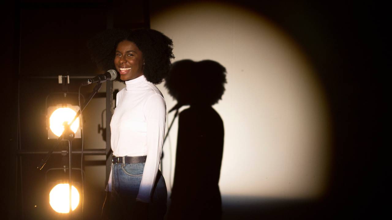 A atriz Indira Nascimento em cena posa em frente a um microfone, com luz de palco, sorrindo