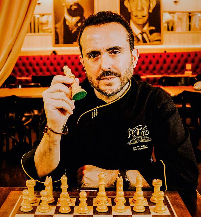 Fundador do Paris 6, Isaac Azar é o vencedor de torneio on-line de xadrez. Na foto, posa sentado no restaurante segurando peça de xadrez com tabuleiro à frente.