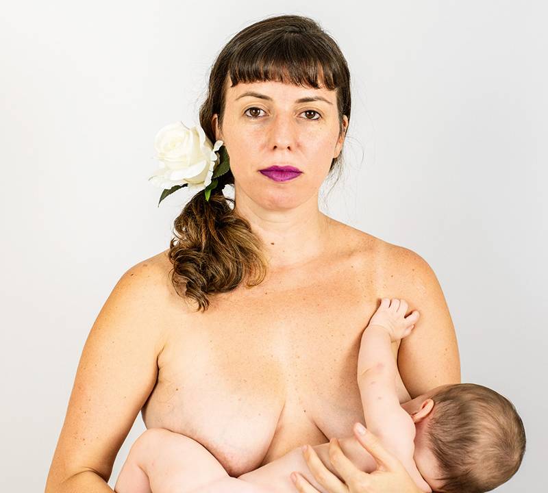 Sem roupas, a mãe e atriz Joana Barbosa segura sua bebê Iara no colo voltada para seu peito direito. Com cabelo preso em rabo de cavalo e batom roxo nos lábios, olha diretamente para a câmera.