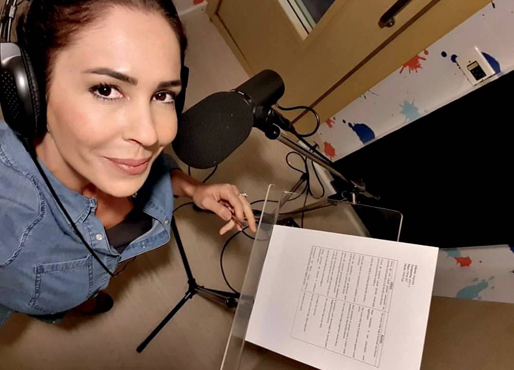 A radialista Juliana Veiga aparece em ângulo visto de cima usando fones de ouvido pretos, vestindo camisa jeans e sorrindo em frente a um microfone profissional e uma folha branca com texto.