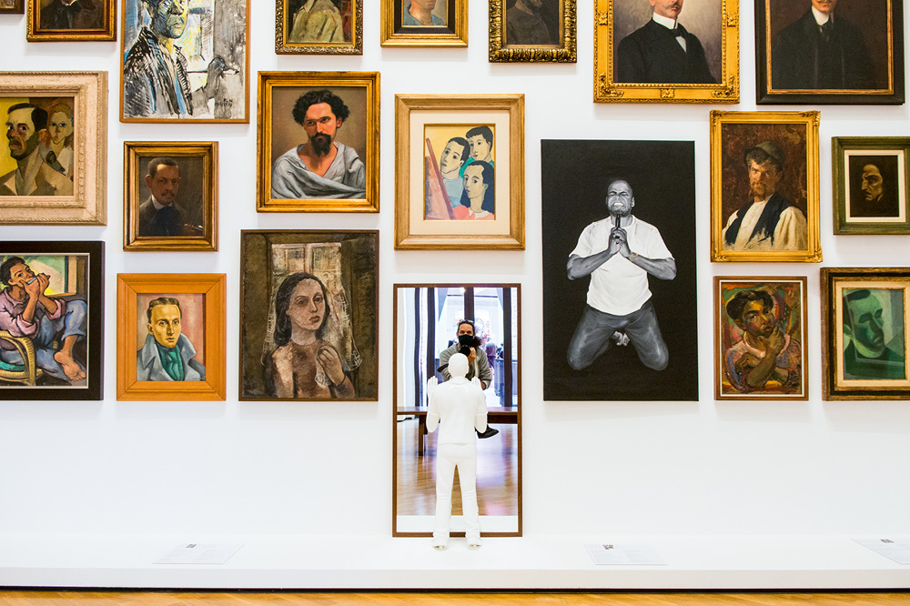 Vários quadros dispostos em uma parede branca, um deles é um espelho e da para ver o fotógrafo tirando a foto