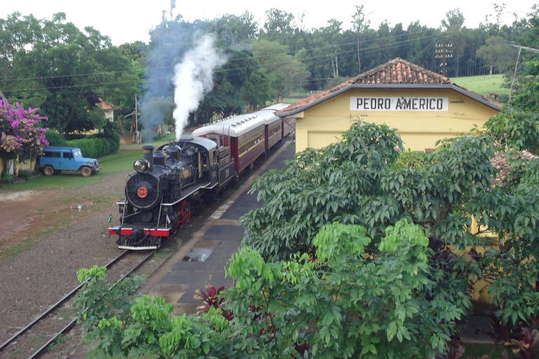 Foto mostra gramado, árvores, estação e maria-fumaça preta e vermelha em rota de Campinas e Jaguariúna.