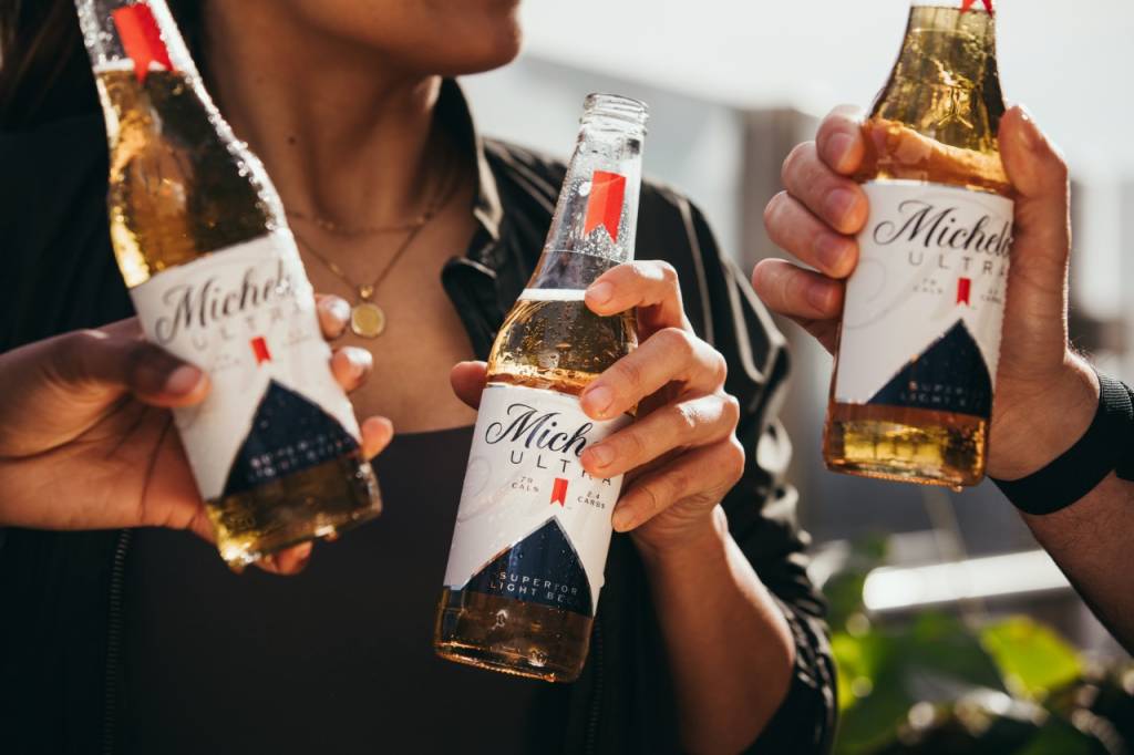 Três pessoas seguram garrafas da cerveja Michelob Ultra, onde o foco da imagem são apenas as mãos e as cervejas