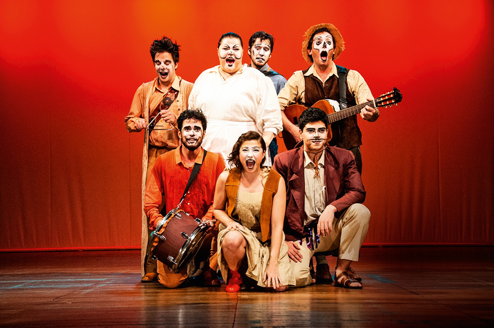 Sete atores do espetáculo O Mágico di Ó estão no palco de um teatro, caracterizados com os rostos pintados de branco. Um segura um tambor e outro um violão