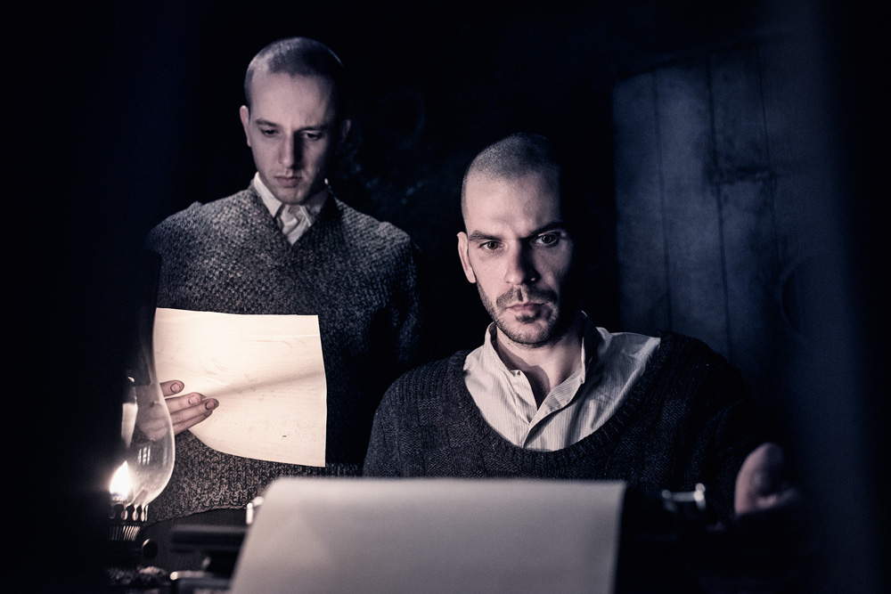 Os dois eslovacos, em uma foto escura. Um deles está em pé com uma folha em mãos e o outro está escrevendo numa máquina de escrever