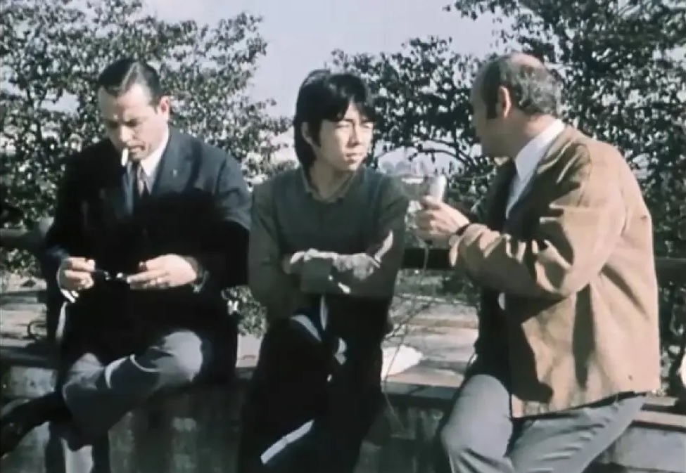 Cena de Os Arrependidos, dirigido por Ricardo Calil e Armando Antenore, mostra três homens sentados na rua conversando. No meio, um rapaz de origem asiática olha para o homem da direita enquanto o da esquerda fuma um cigarro.