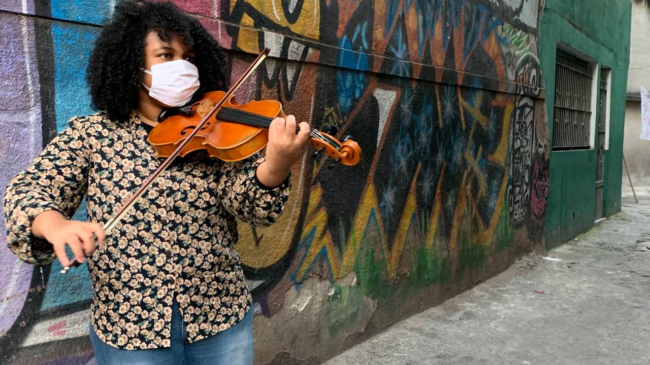 Estudante Palloma toca viola erudita de pé em frente a paredão colorido.