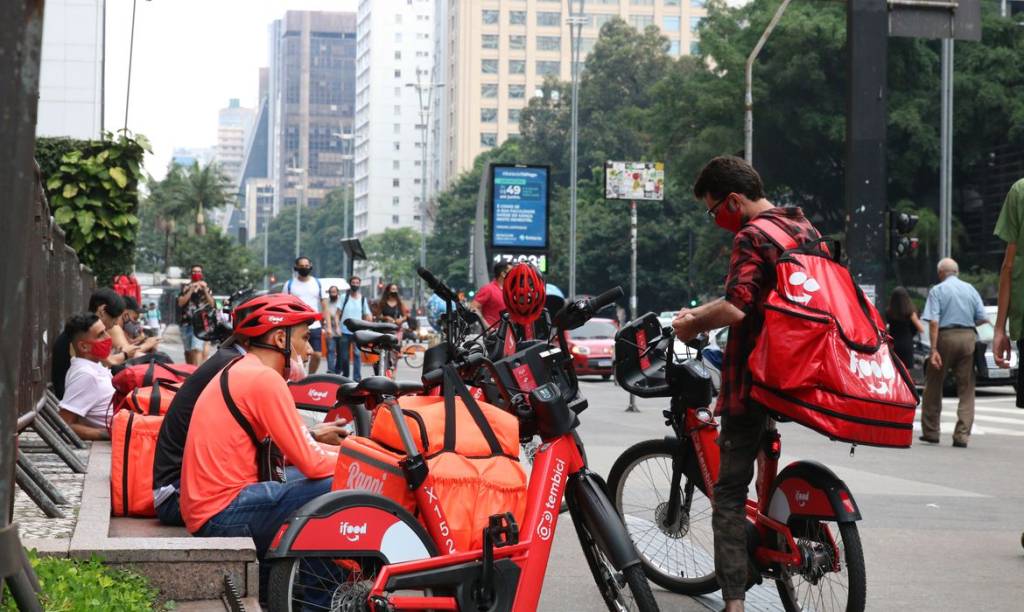 Imagem na Avenida Paulista mostra entregadores sentados e parados na calçada com bicicletas e roupa laranja da marca Rappi.