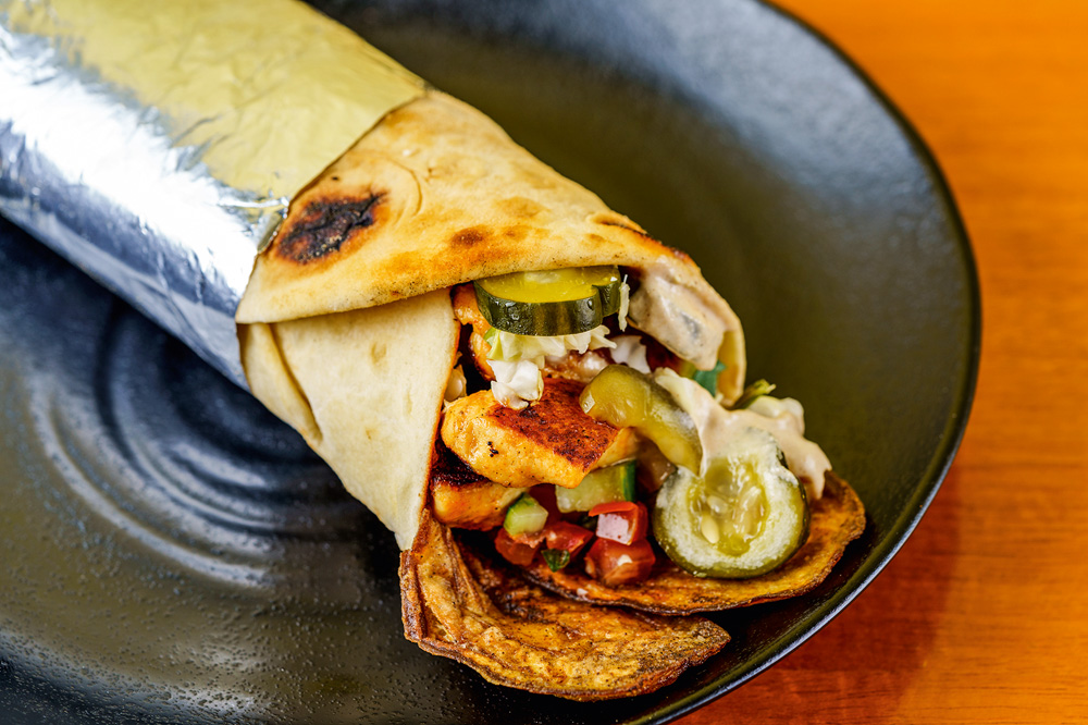 Sanduíche enrolado, similar a um kebab, sobre prato de cerâmica com pedaço de frango, pepino, molho tahine e berijenla frita aparecendo.