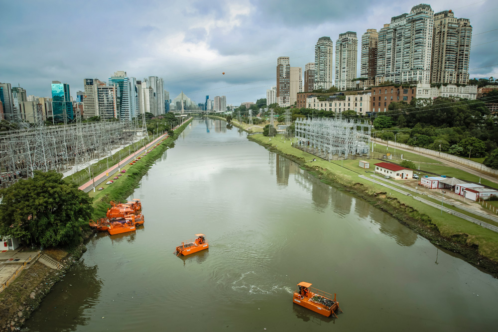 Imagem do Rio Pinheiros visto de longe. É possível ver pequenas embarcações laranjas em baixo, margens e prédios do entorno