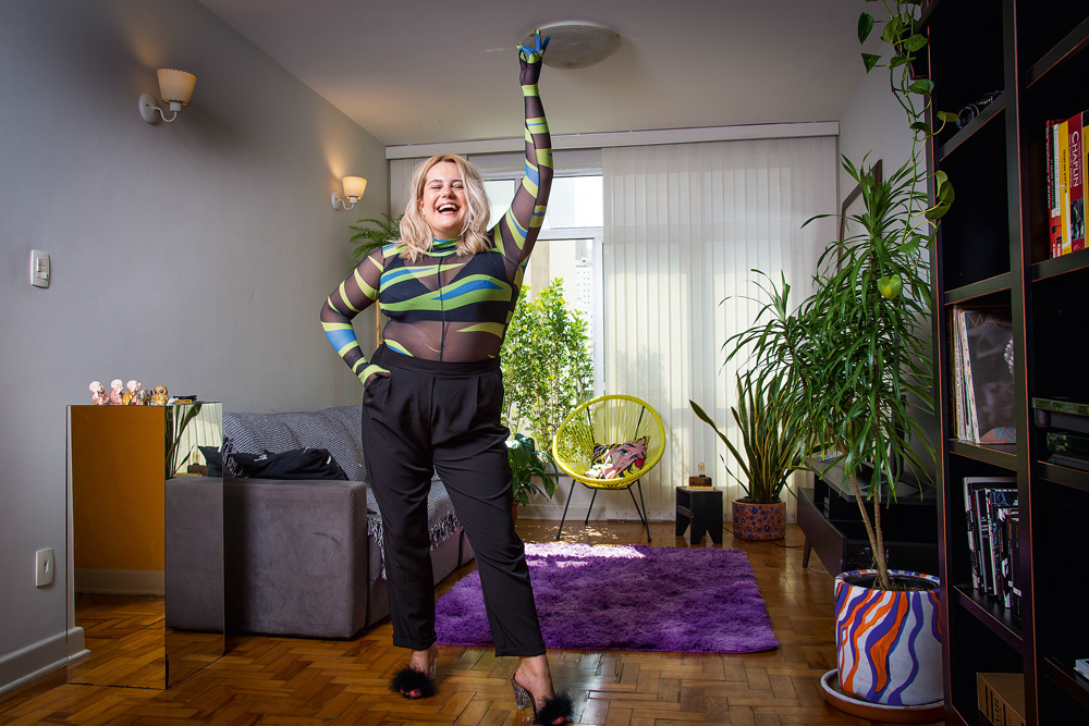 Luiza Gil, fundadora da A.rolê, fazendo pose para a foto na sala de seu apartamento com uma mãe no bolso e a outra apontando para cima