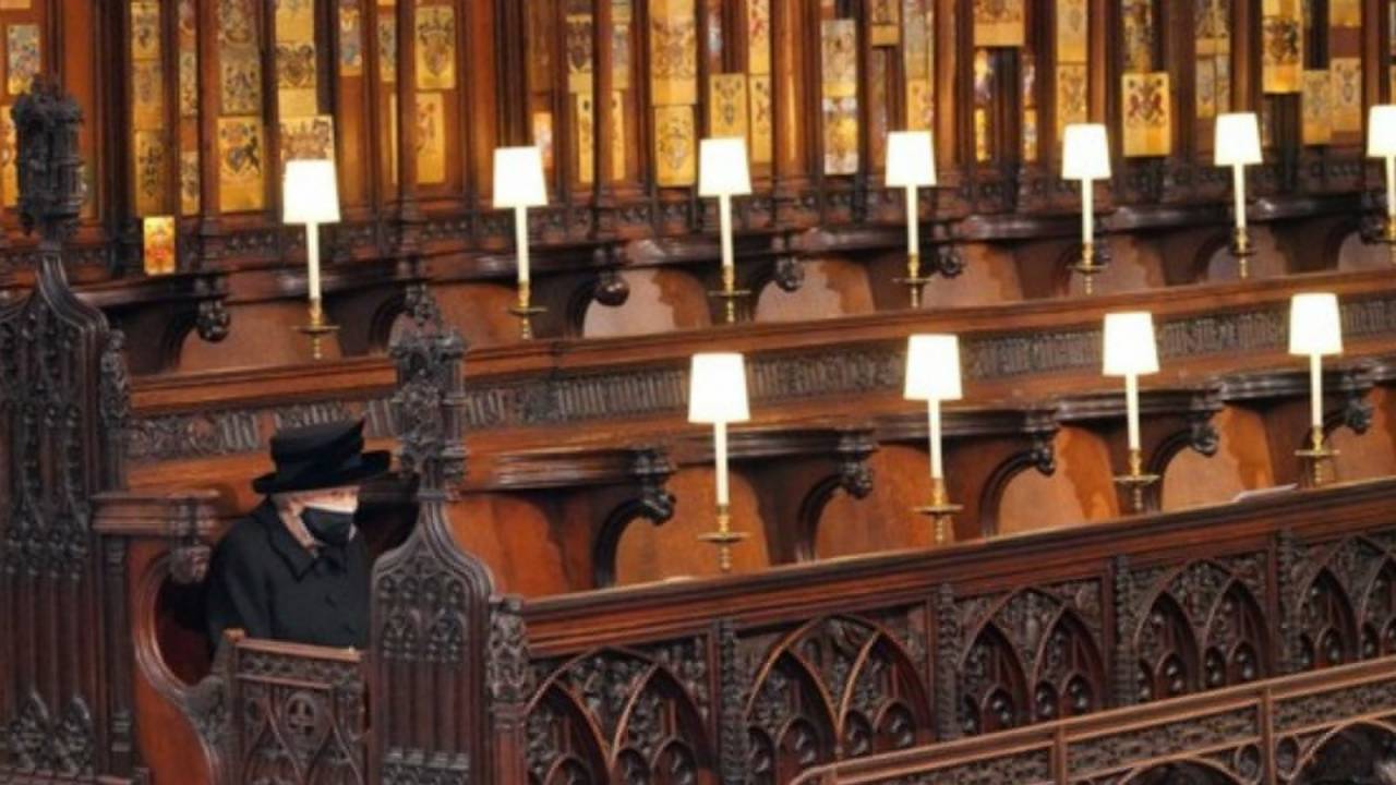 Rainha Elizabeth II aparece sentada sozinha em fileira de cadeiras de madeira com luminárias individuais. Veste roupas pretas para funeral do marido.