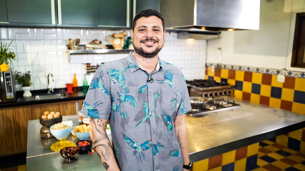Raul Lemos, de pé, sorrindo para a foto encostado em uma bancada de cozinha, atrás prateleiras com utensílios e fogão do lado direito.