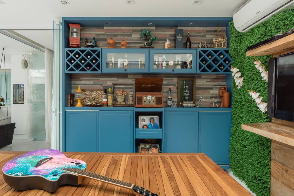 Móveis na cor azul e mesa de madeira de demolição ocupam espaço da casa reformada do músico Marcos. Na mesa, aparece uma guitarra colorida.