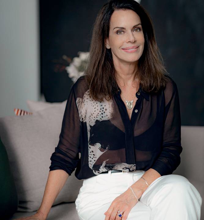 A empresária e influencer Renata Scarpa posa com calça branca e camisa preta transparente. Aparece na foto sorrindo, de pernas cruzadas e com uma das mãos encostada no assento.