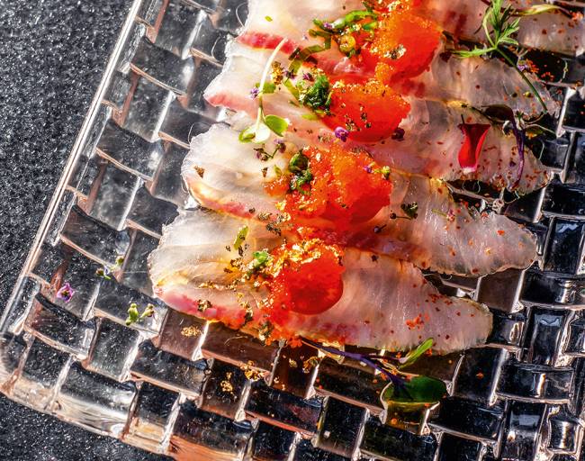 Prato retangular transparente com fatias de peixe finas cobertas por ovas de peixe e pimenta.