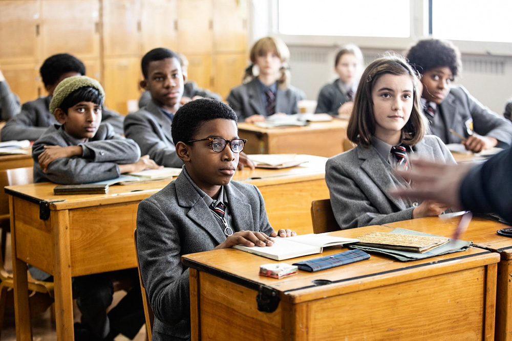 O personagem Kingsley no centro de imagem, dentro de uma sala de aula com outros colegas. Todos estão sentados e olhando para frente