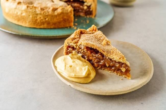 Pedaço de torta de maçã sobre prato de cerâmica ao lado de creme amarelado. Ao fundo, a torta inteira sem o pedaço à frente.