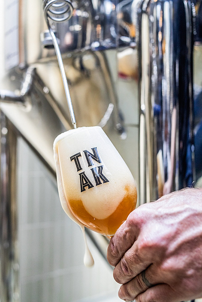 Pessoa se servindo da cerveja direto do barril. Na imagem aparece uma mão da cor branca, com a taça de vidro com o logo do tank, enquanto a cerveja sai do barril.