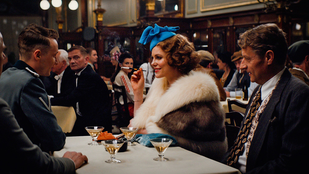 Cena do filme A Espiã, em que a protagonista está em uma mesa de um restaurante com outros homens