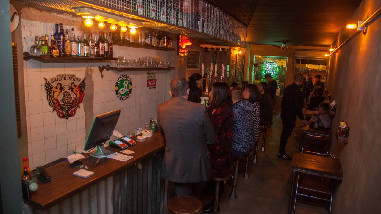 Ambiente de bar com luz baixa, balcão com banquetas e pessoas sentadas à esquerda, mesinhas à direita.