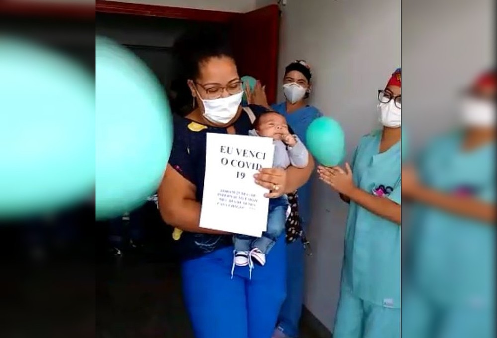 mãe mical segurando o bebê luís miguel, sendo aplaudida por médicos e com uma folha com 'eu venci a covid-19' escrito