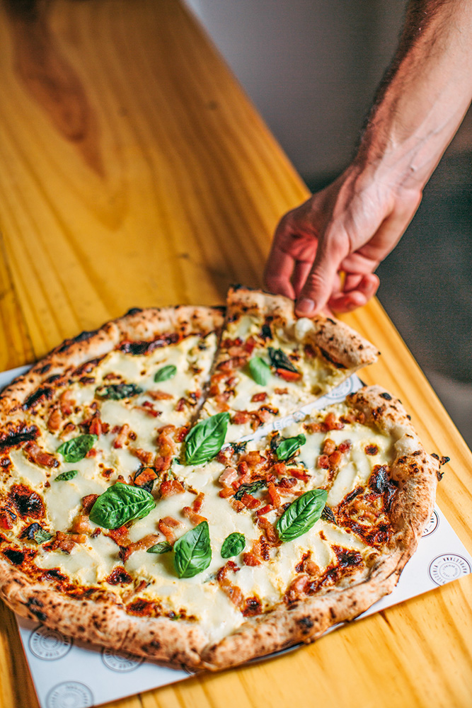 Ciao Pizzaria Napoletana – Pizza Pessoa retirando uma fatia da pizza que esta sobre balcão de madeira