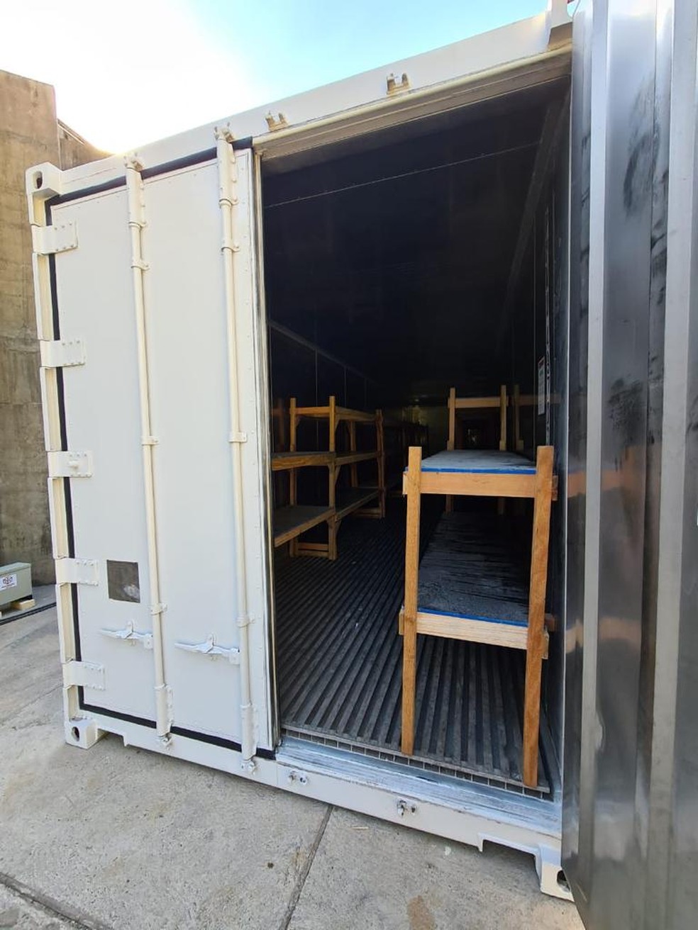 Imagem mostra contêiner com uma das portas abertas. Dentro dele, beliches para guardar os corpos