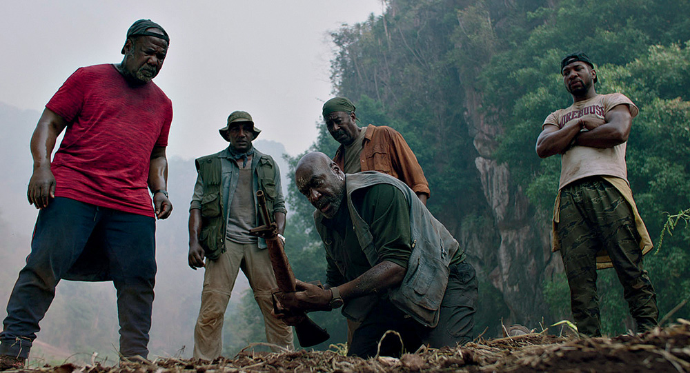 Cinco homens negros olham para um objeto segurado por um deles perto do chão de terra