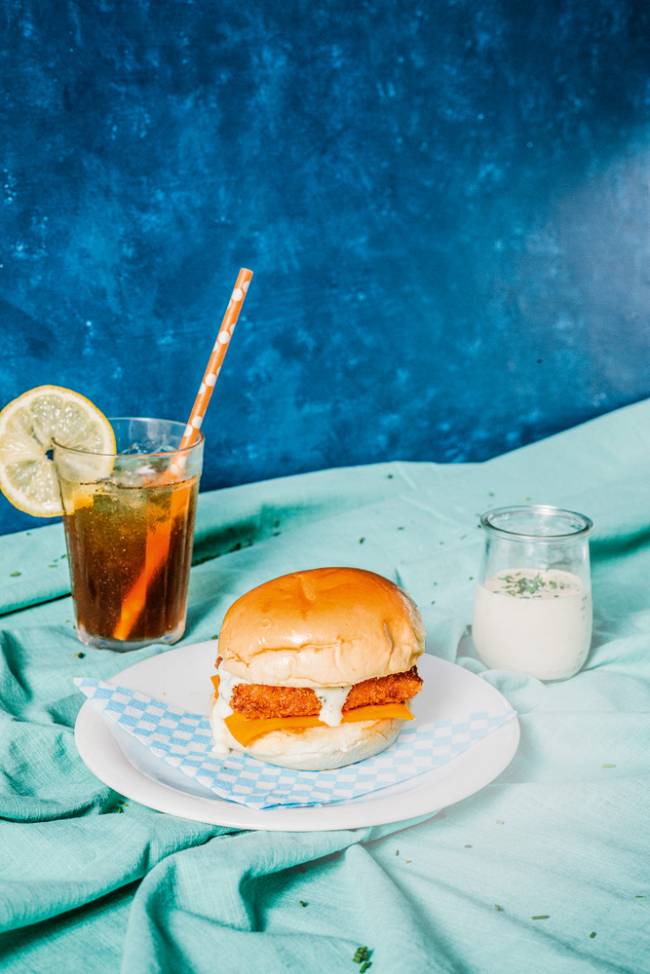 hambúrguer de peixe sobre prato com copo de mate com limão do lado esquerdo e pote de maionese do lado direito