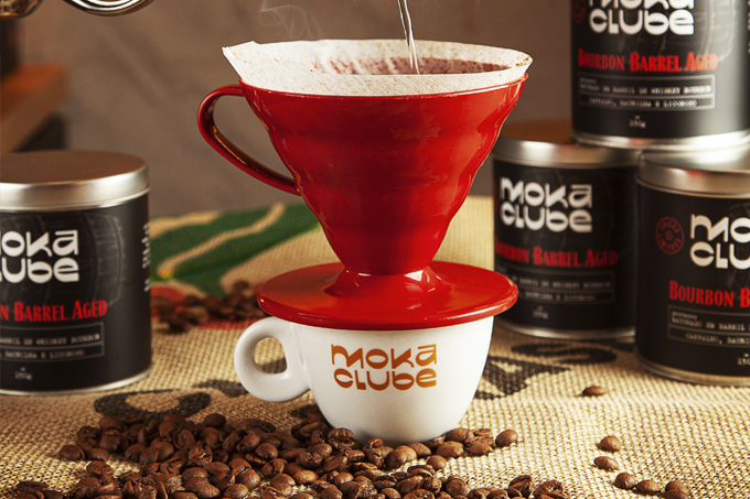 Xícara de café branca com logo em vermelho ao centro, cercada de grãos de café, sob coador vermelho de café que recebe água. Ao fundo, latas pretas de metal de café com logo branco e vermelho.