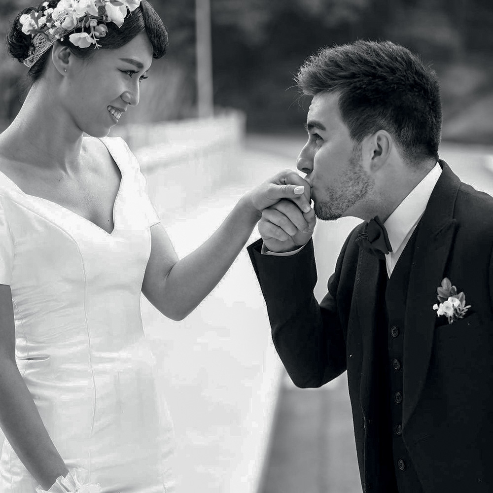 Casal vestido de noivo e noiva, de lado para a foto, com Lucas beijando a mão de Sisi
