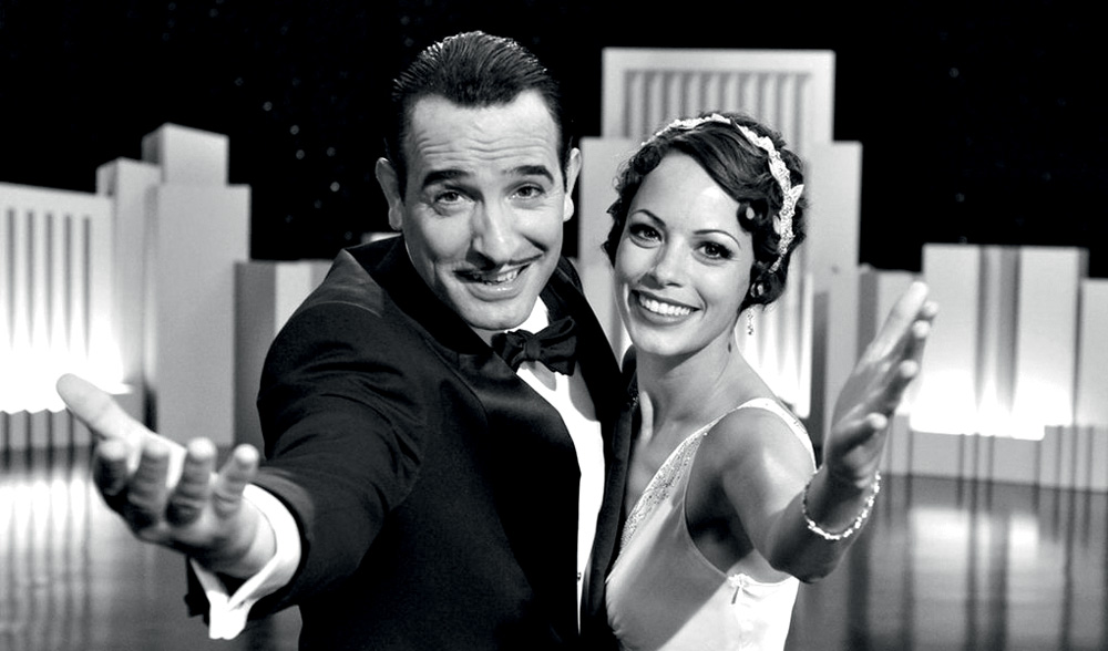 Em preto e branco, um homem e uma mulher, lado a lado, esticam o braço para câmera sorrindo