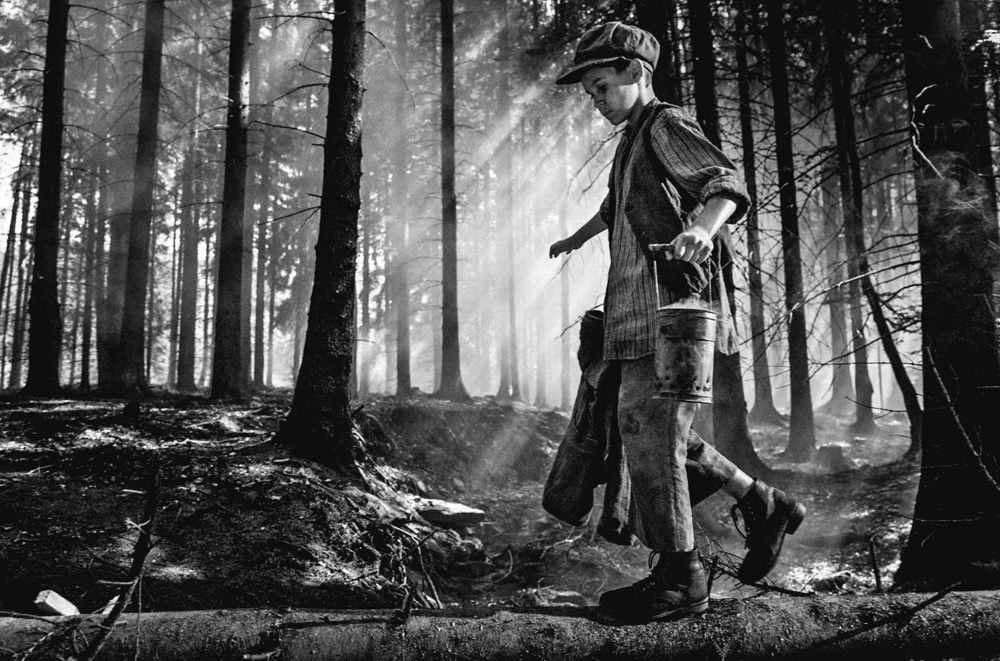 Petr Kotlár, em uma imagem preta e branca, anda com roupas tradicionais do leste europeu da época da ll Guerra. Ele está em meio a uma floresta.