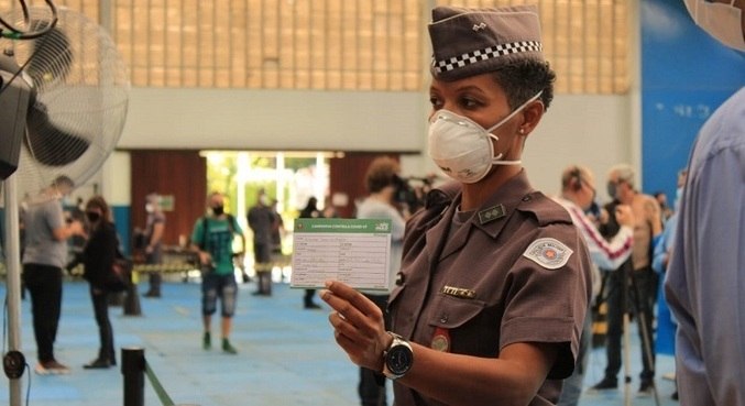 Tenente, de uniforme marrom e máscara, mostra papel de vacinação