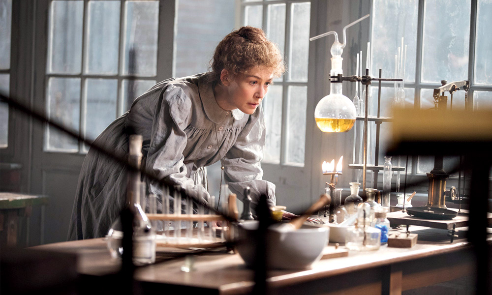 Rosamund Pike no papel de Marie Curie em frente a uma mesa com experimentos químicos